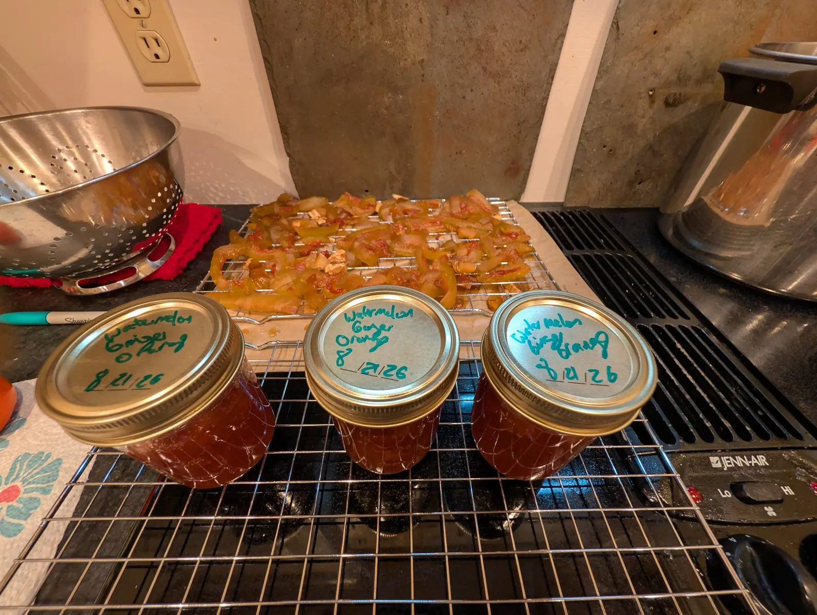 Three half-pints of amber preserve cooling on the stovetop rack, hand-dated on the lid, and behind them the candied peel still draining — each strip translucent now, pale green at one edge and blushed pink at the other.
