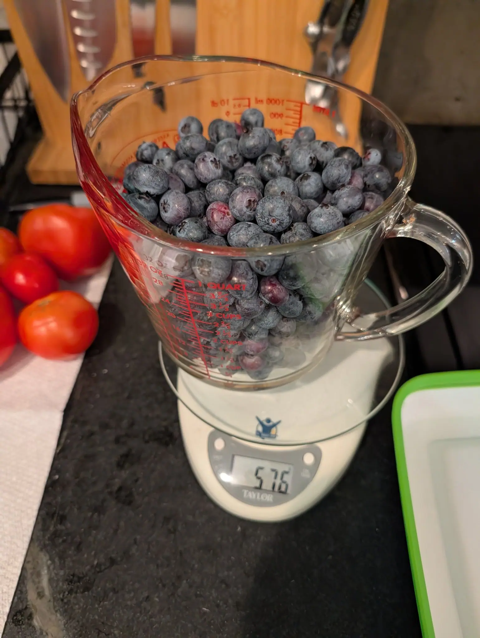High Bush blueberries on 7 August, heaped in a glass pitcher until the top ones stand above the rim, gray bloom thick over purple-black skin and fine russet lines scratched across a few — one in the front row still pink-red and unripe.