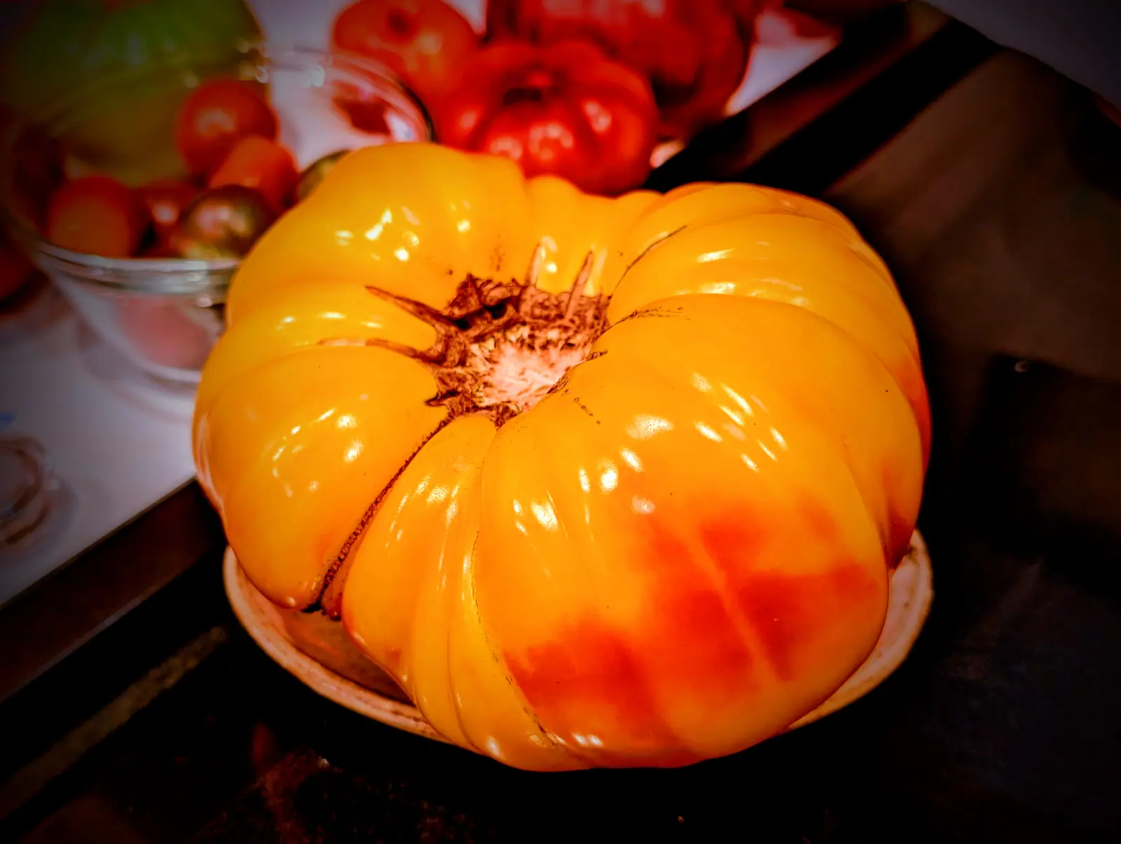 A Pineapple Tomato on 25 August, deeply ribbed and glossy yellow-orange with its one red blush low on a shoulder rather than at the top, set alone on a small saucer with a bowl of smaller fruit behind it and the frame darkening at its edges.