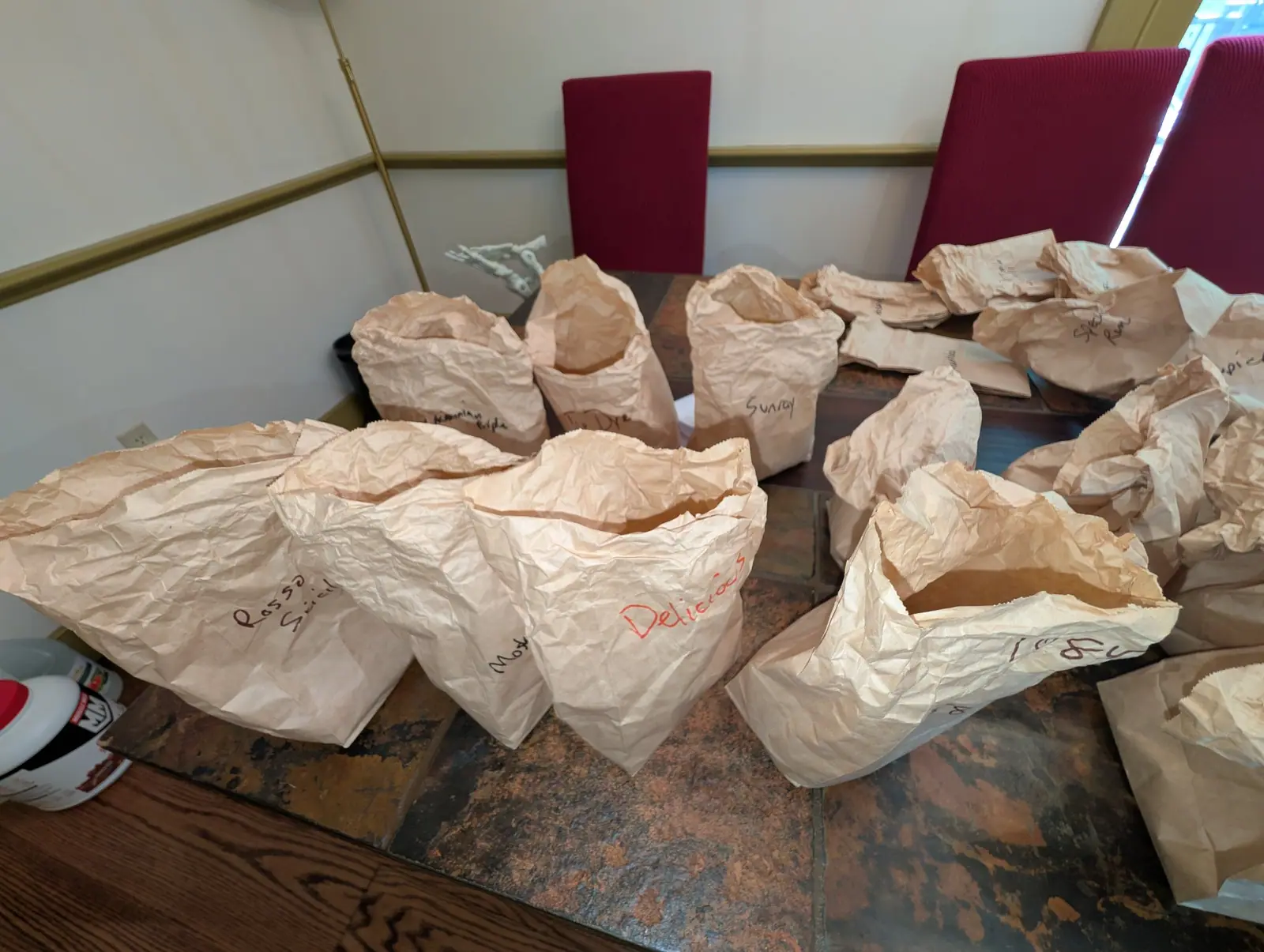 About twenty brown paper bags stand open across a slate tabletop on 12 August, every top flared wide and rolled back on itself so the insides read as dark cavities, cool window light from the right lifting their pinked edges bright against the stone.