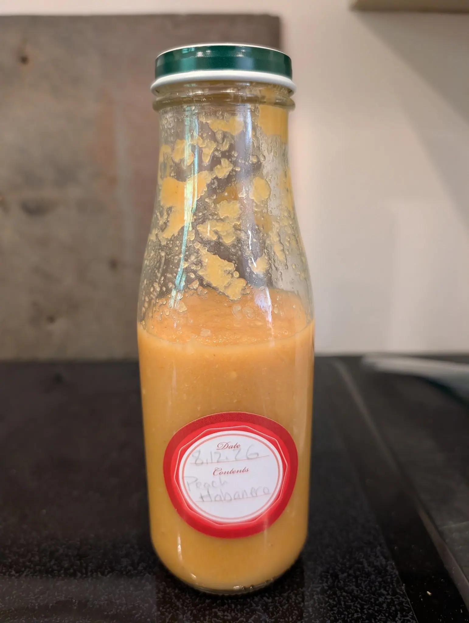 A tall glass bottle alone on a dark counter on 12 August, filled a little past halfway with an opaque peach-orange puree under a band of fine bubbles, the glass above the fill line spattered with gobs and runs of it and beaded with condensation.