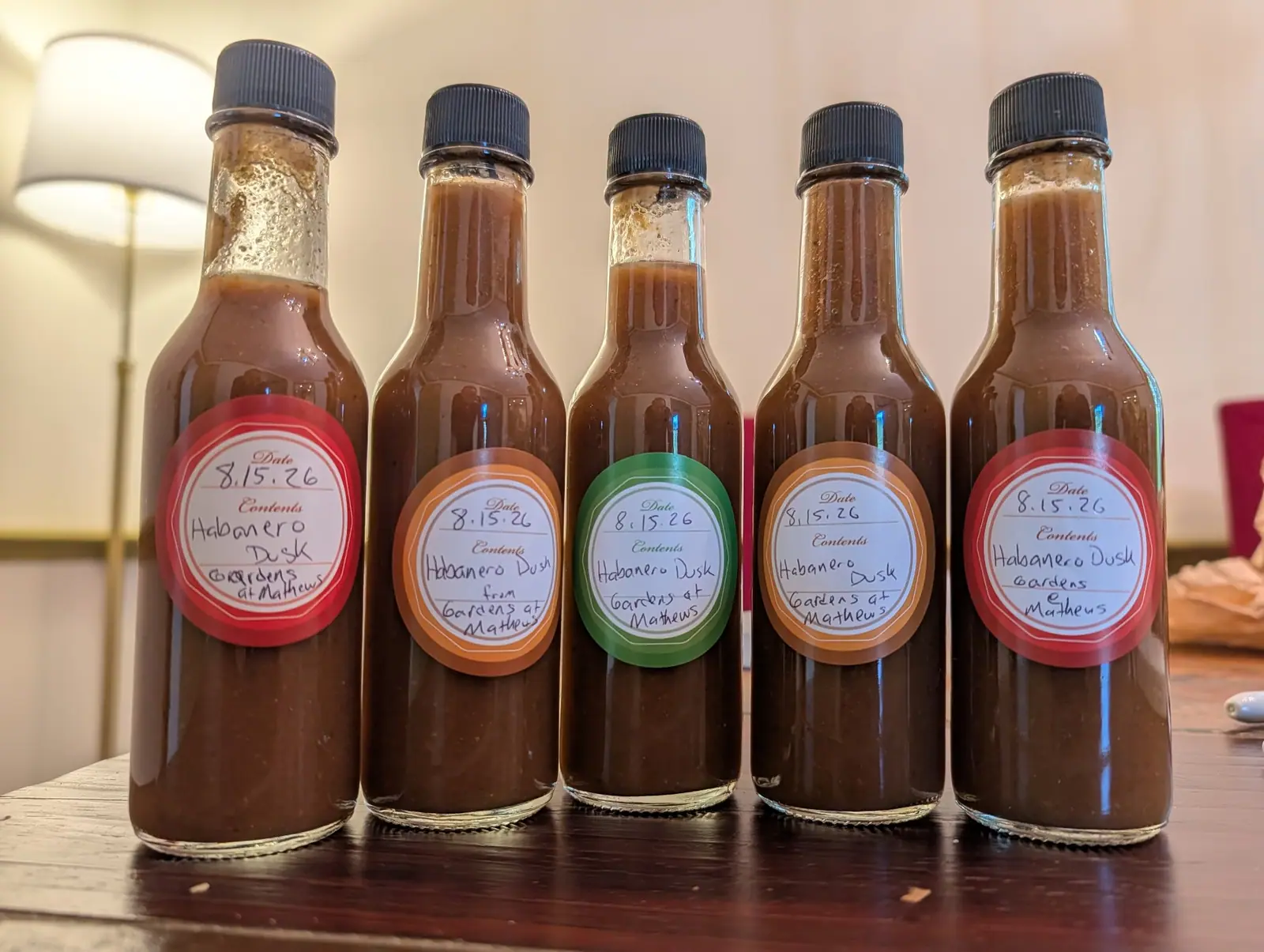 Five bottles of thick red-brown sauce in a row on dark wood on 15 August, the first and third filled only to the shoulder with condensation beading the empty neck glass. Lamplight from the left rakes a single highlight down every shoulder.
