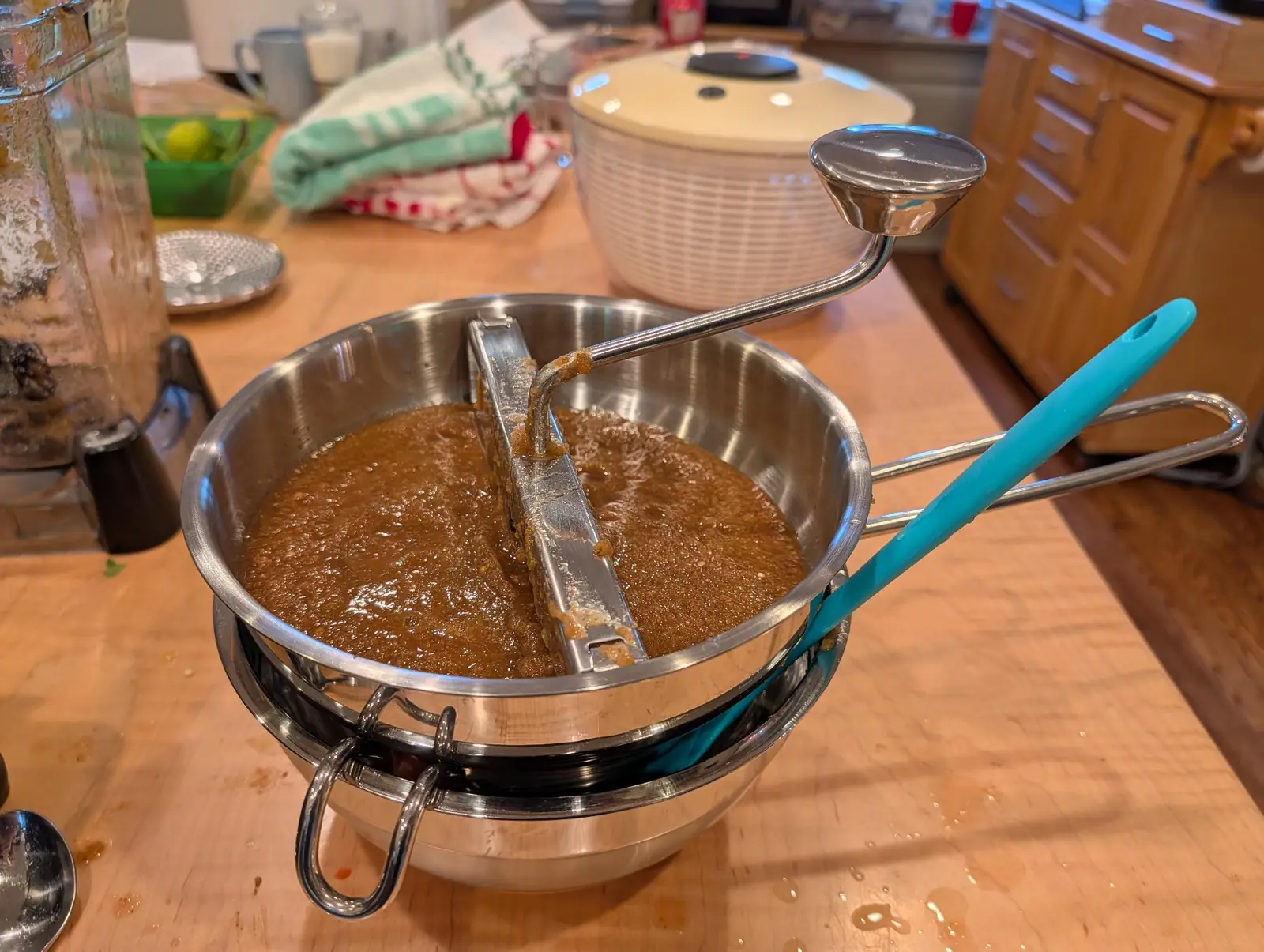 A rotary food mill sunk into the rim of a mixing bowl on 15 August, two-thirds full of orange-brown puree gone matte and frothed at the surface, a clot of pale pulp caked around the crank hinge. Amber juice has dried in splashes across the butcher block.