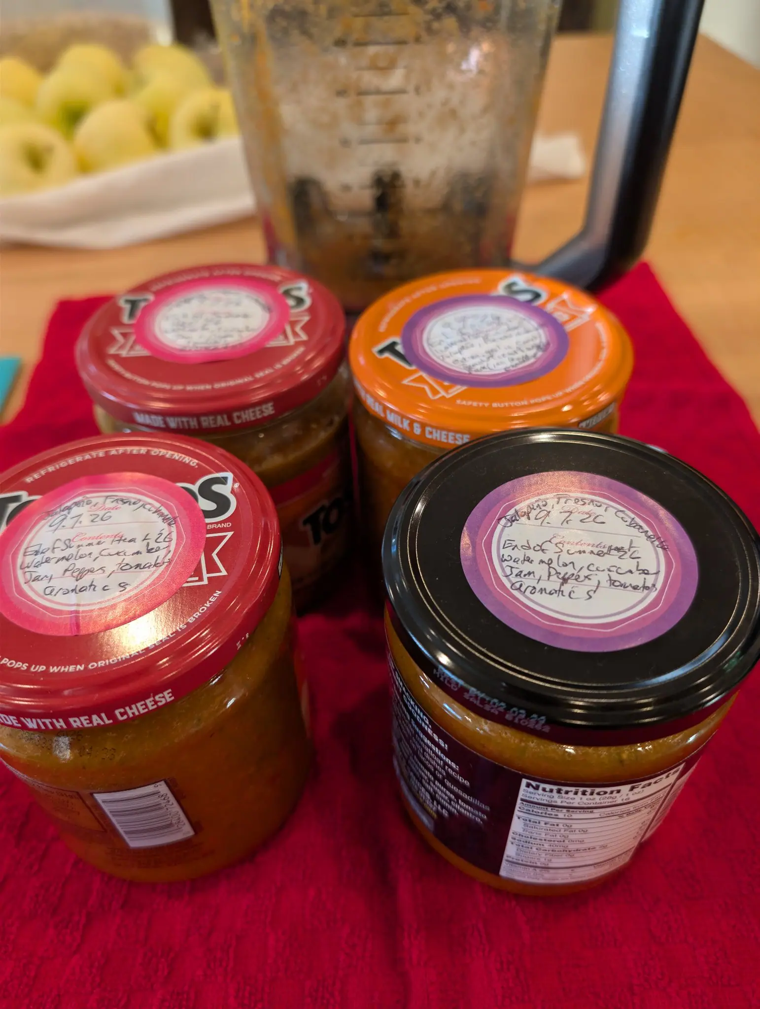 Four re-used jars on a red cloth on 8 September, each packed to the shoulder with a coarse orange-brown relish flecked green and dark, their lids mismatched — two red, one orange, one black — and every one carrying a hand-written sticker.