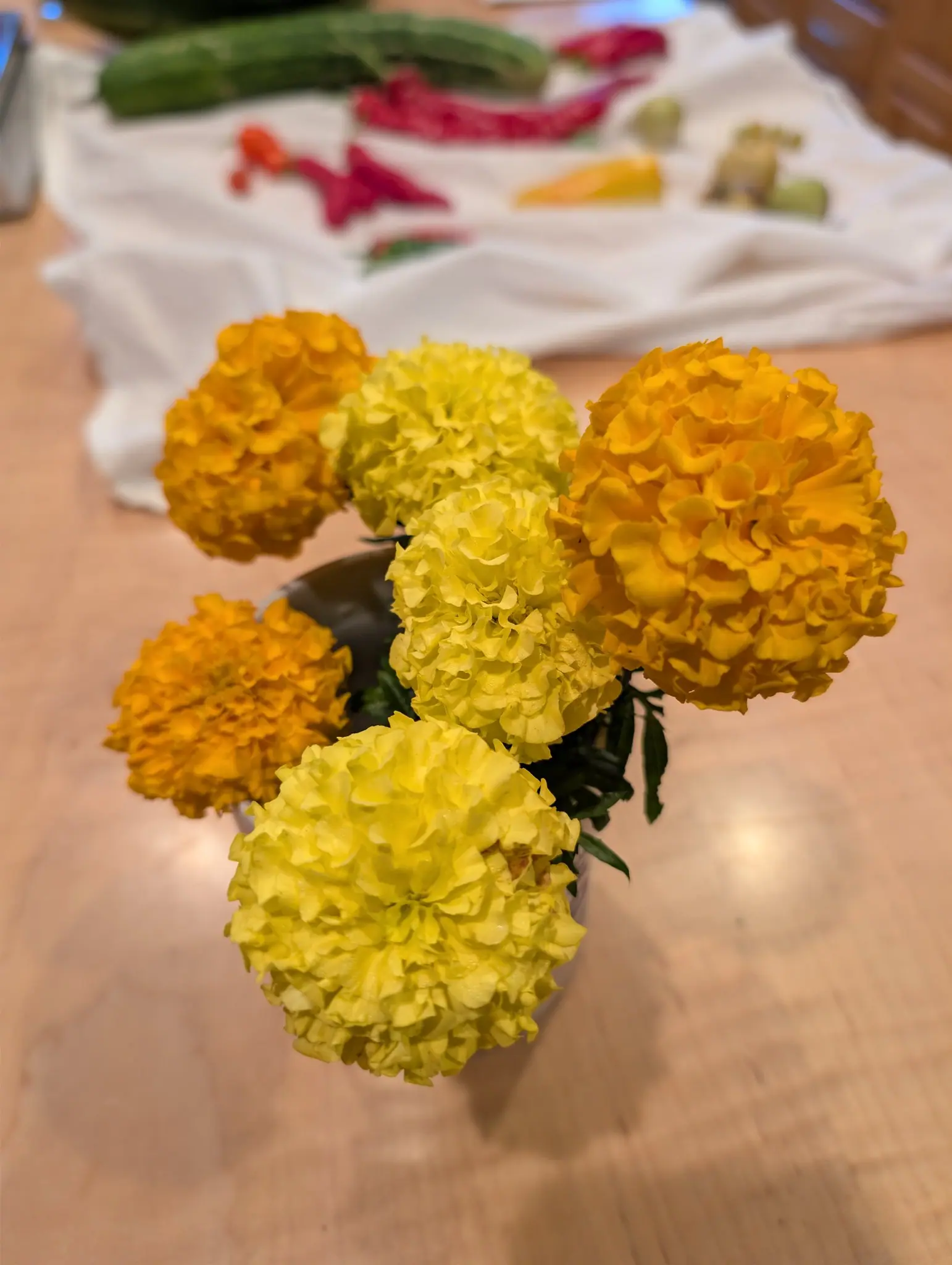 A cut bouquet of six marigolds on a blond wood table on 11 September, three deep golden orange and three lemon yellow set alternately, each a dense ball of ruffled, crinkled florets — a harvest spread blurred on a white cloth behind, in warm indoor light.