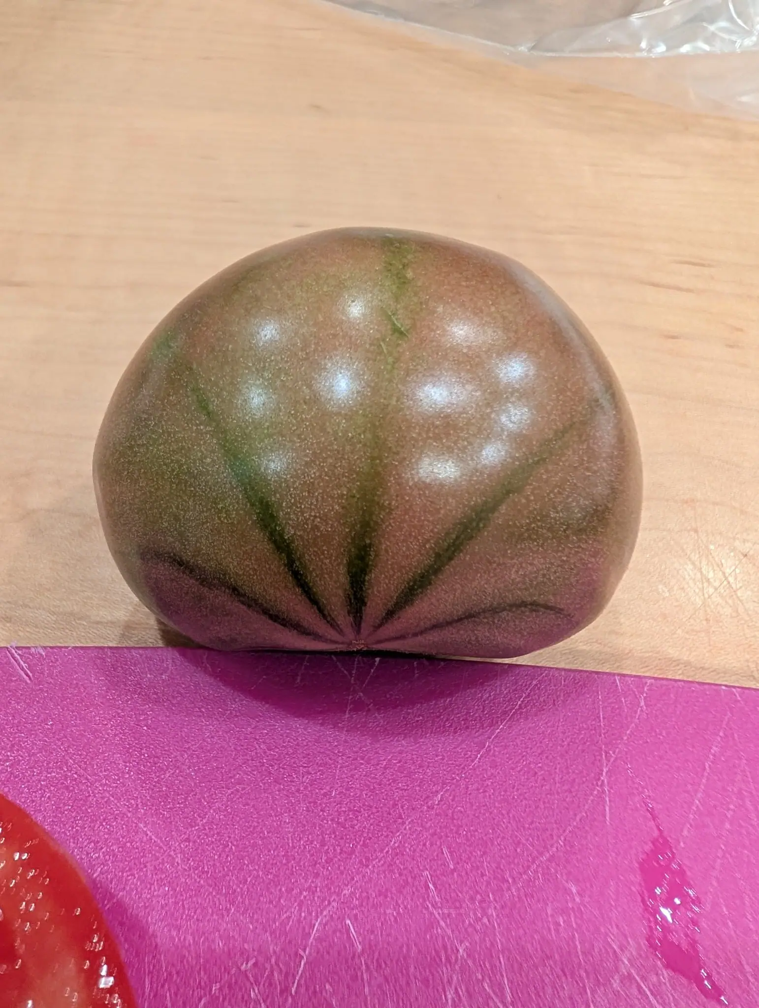A Manitoba tomato stands upright on its blossom end on 14 September, six or seven near-black lines fanning from the pale blossom scar across dusky rose and green shoulders. Flat kitchen light; a cut half lies on the magenta board below.