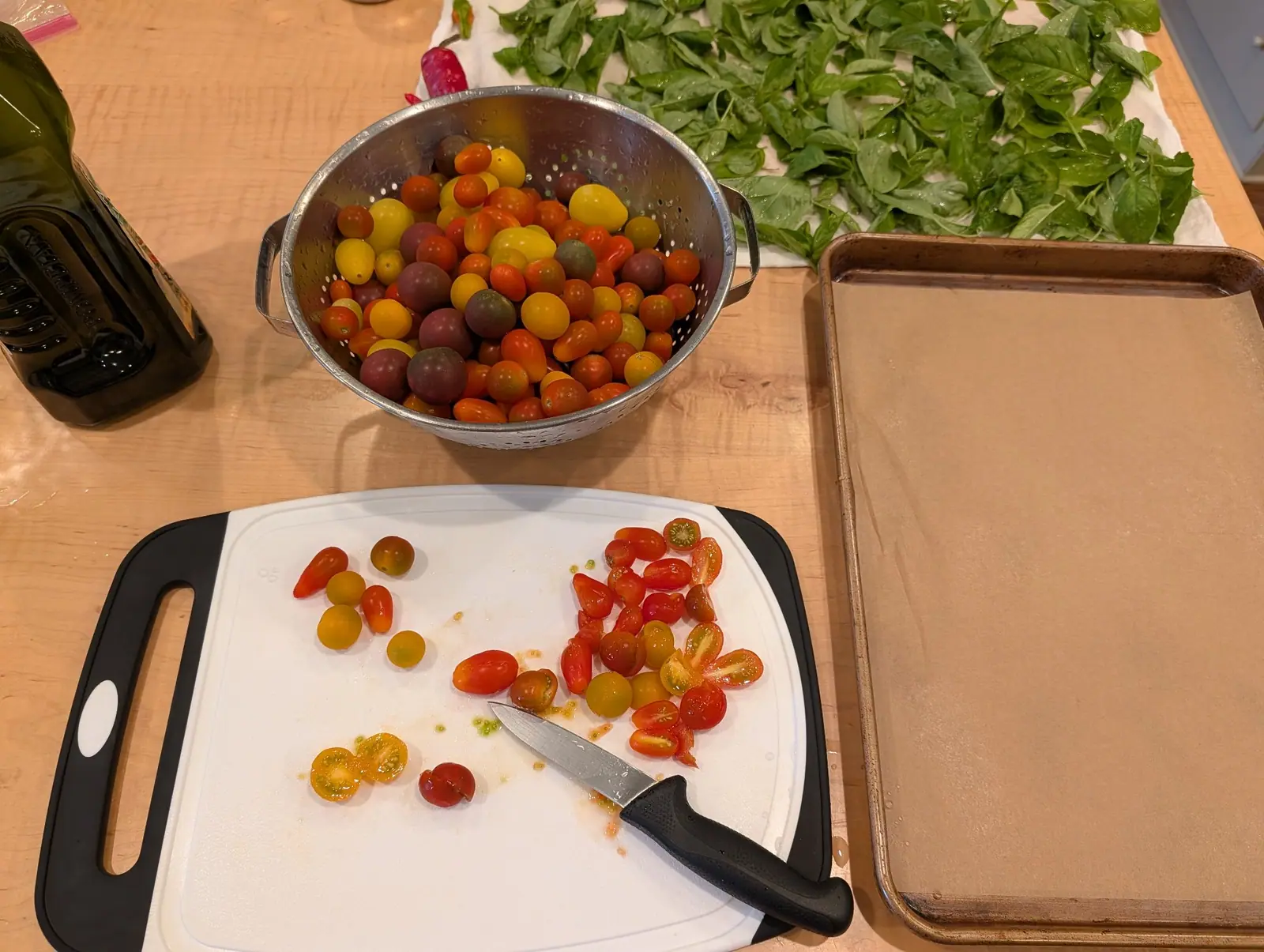 Washed fruit sorted on 16 September: scarlet, yellow, and dusky near-black globes heaped in a dented colander, about forty already halved cut side up on the board, seed cavities glistening. Warm overhead light, no daylight.