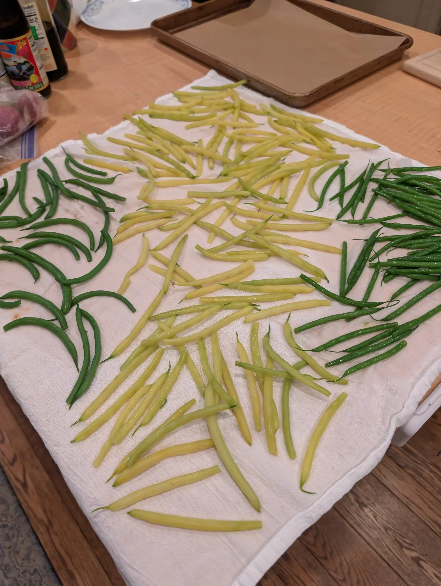 Washed pods drain on a flour-sack towel on 22 September, the pale yellow ones in the center still carrying a faint green line along the suture and a fine curled tip at the tail, slim dark green arcs flanking them. Warm kitchen light, no daylight.