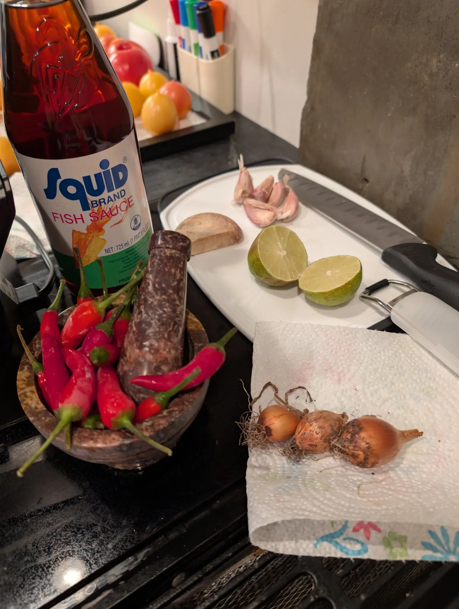 Chili Red peppers on 7 October, heaped in a stone mortar with the pestle standing upright among them, most a glossy scarlet but one at the center still almost wholly green; hard indoor light at night rakes the fruit and the background falls away dark.