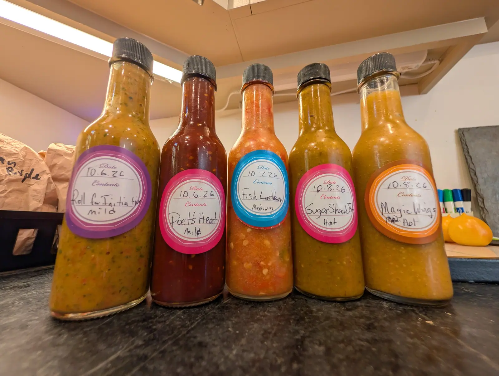 Five woozy bottles in a row on mottled gray stone on 8 October, filled nearly to the neck and ranging from an opaque olive puree flecked with seeds to a loose orange-red sauce with pale seeds settled through it. Hard tube light from above crosses the caps.