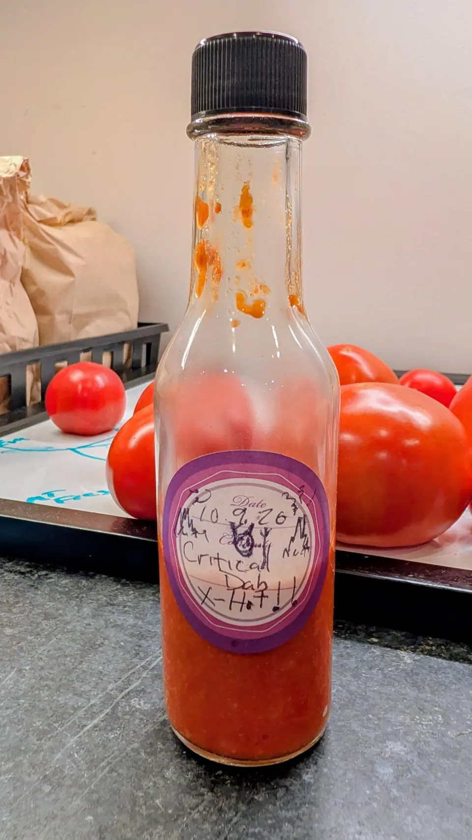 A woozy bottle of opaque orange-red sauce on 9 October, filled to two thirds with pale flecks and seeds suspended in it, the empty neck above the fill line streaked with dried splashes; soft indoor light, a blurred row of glossy red fruit behind.