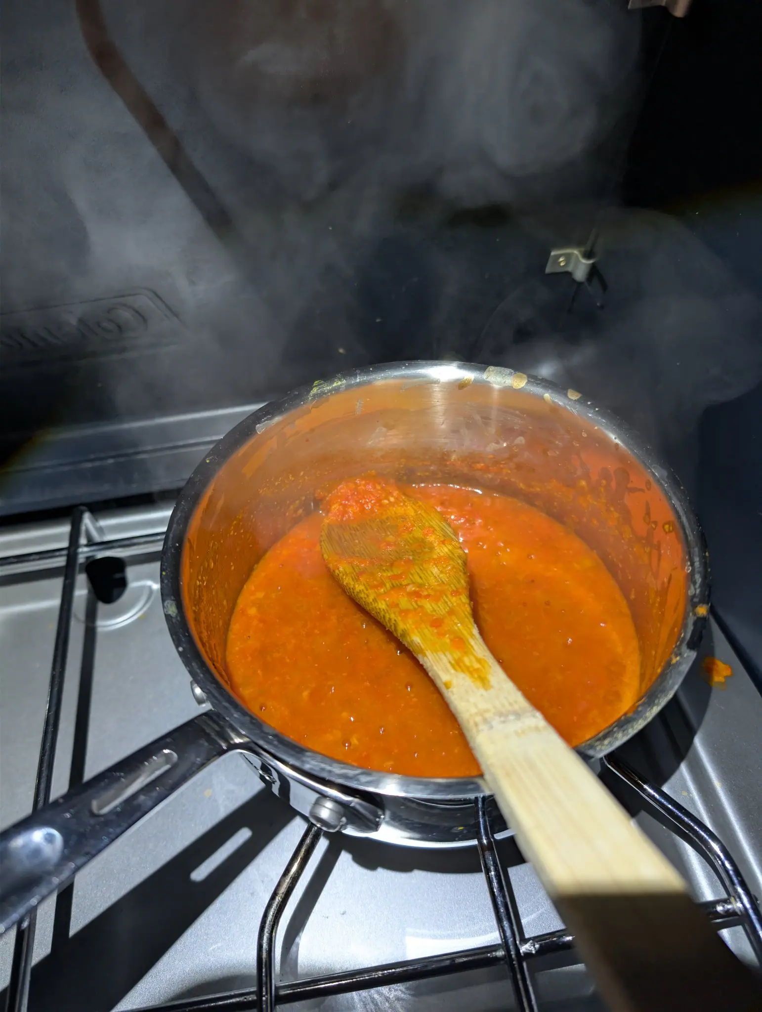 A coarse orange-red puree simmers in a stainless saucepan on 9 October, tiny seeds and paler flecks suspended in the gloss, the wooden spoon's bowl half-submerged and stained dark with clinging pulp. Hard front light leaves the panel behind in shadow.