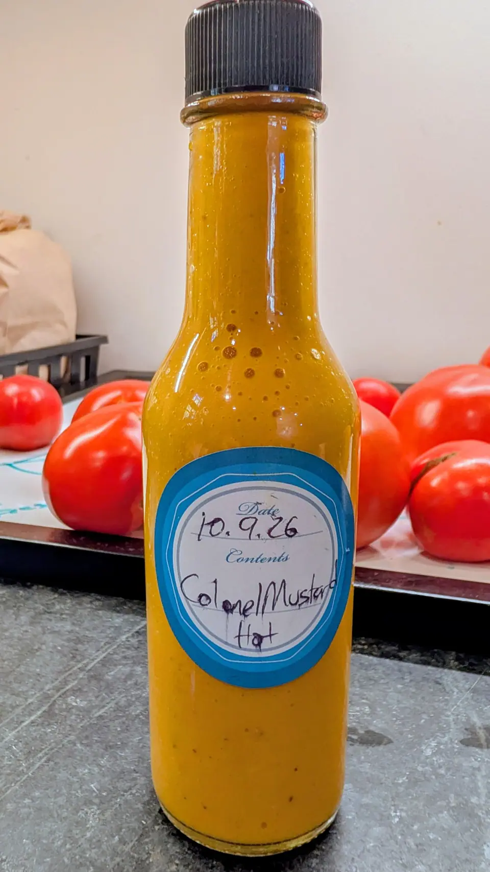 Banana Creme tomatoes put up as a thick opaque golden-yellow sauce, upright in a tall glass bottle on 9 October with small bubbles collected under the shoulder of the neck; at least six glossy red-orange fruits lie behind in soft diffuse indoor light.