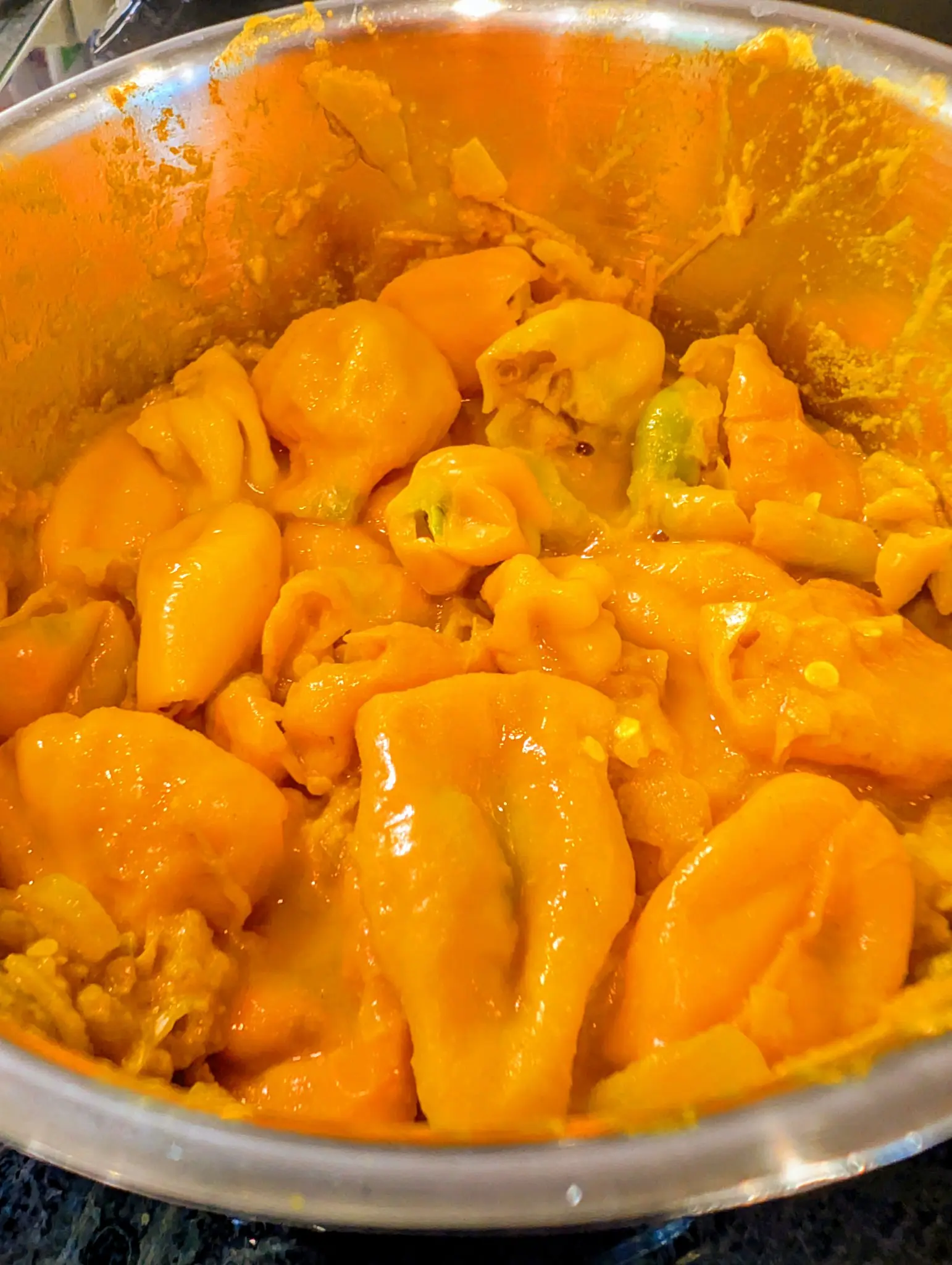 Fifteen or twenty soft orange-yellow fruits heaped in a steel mixing bowl on 9 October, skins slipped into pleated folds, a few green calyx ends and pale cream seeds showing; orange pulp streaks the walls above the pile under warm artificial light.