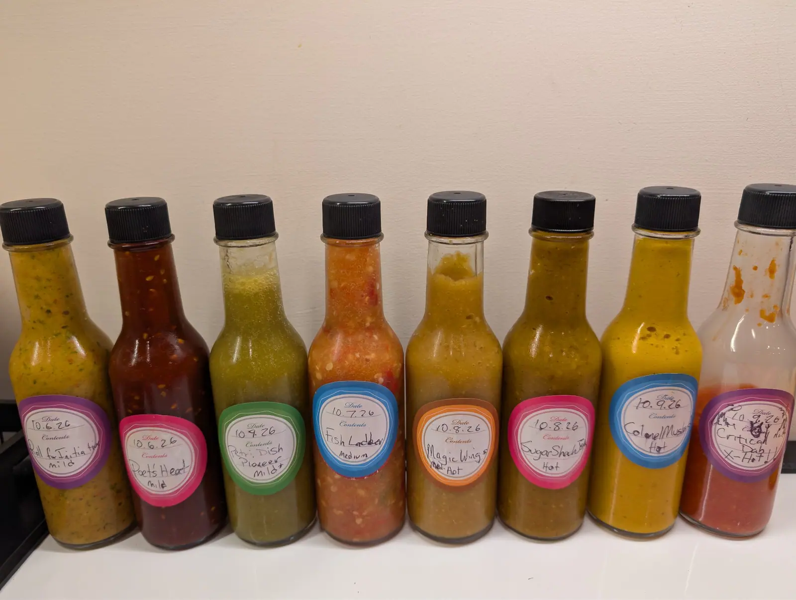 Eight woozy bottles stand shoulder to shoulder in one row on 9 October, each with a black ribbed screw cap; the fourth from the left has separated into a coarse orange-red mash above a paler settled layer. Flat indoor light throws soft shadows to the right.