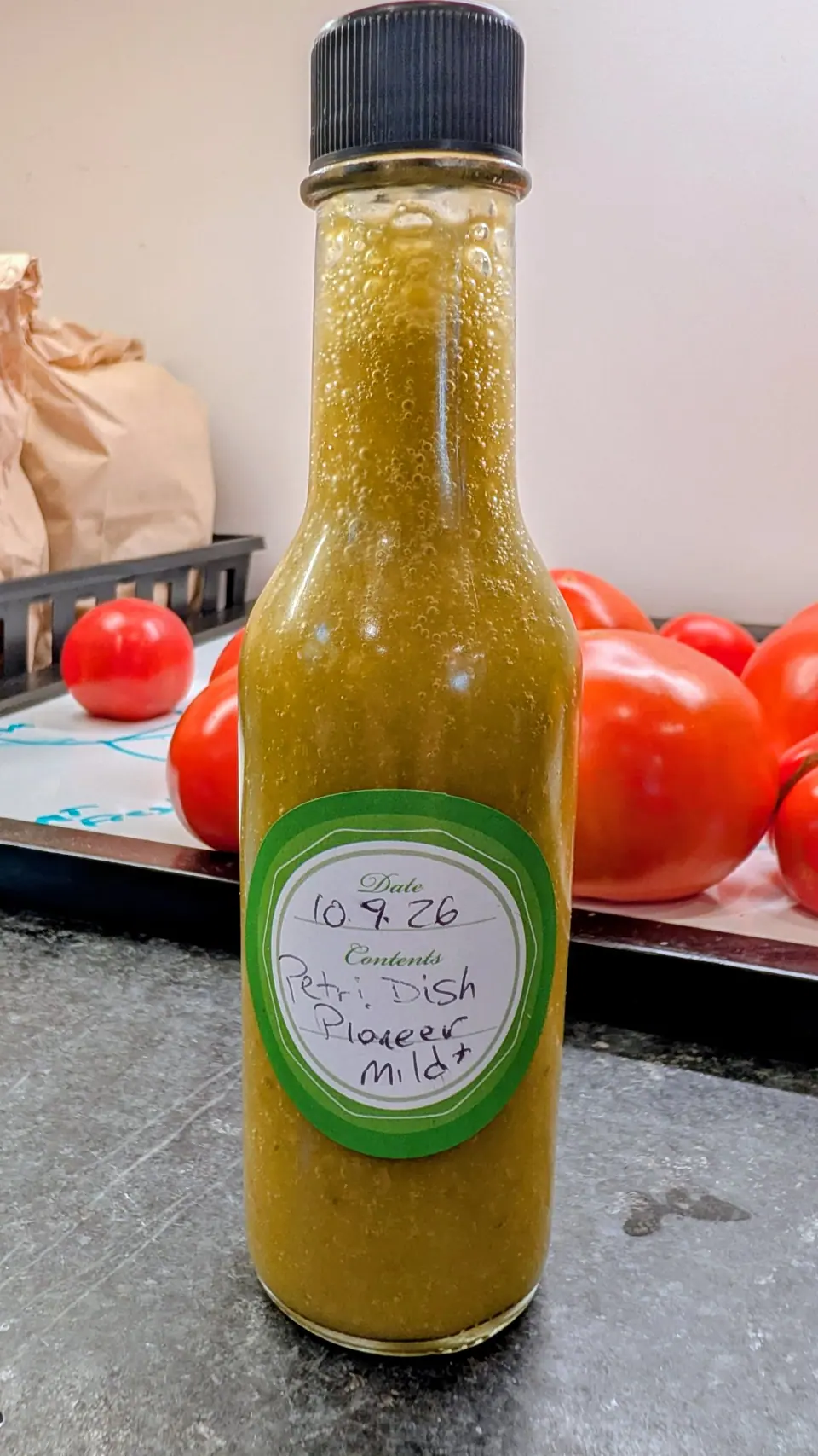 A capped glass bottle stands on gray speckled stone on 9 October, filled to the shoulder with thick mustard-olive puree flecked with darker green and a band of fine bubbles trapped at the neck. Behind it a steel pan of glossy red fruit, flat indoor light.