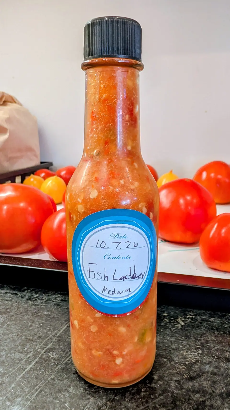 On 8 October, a small clear woozy bottle stands on a dark speckled counter, filled to the shoulder with a coarse orange-red ferment: pale flat seeds and fine bubbles held against the glass just under the black ribbed cap, in soft flat indoor light.
