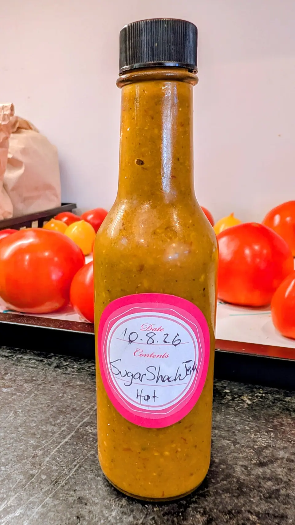 A tall narrow bottle stands capped on a dark speckled counter on 8 October, filled to the shoulder with opaque mustard-amber puree, thick and grainy with seeds and flecks suspended against the glass; flat overhead light, a tray of glossy red fruit behind.