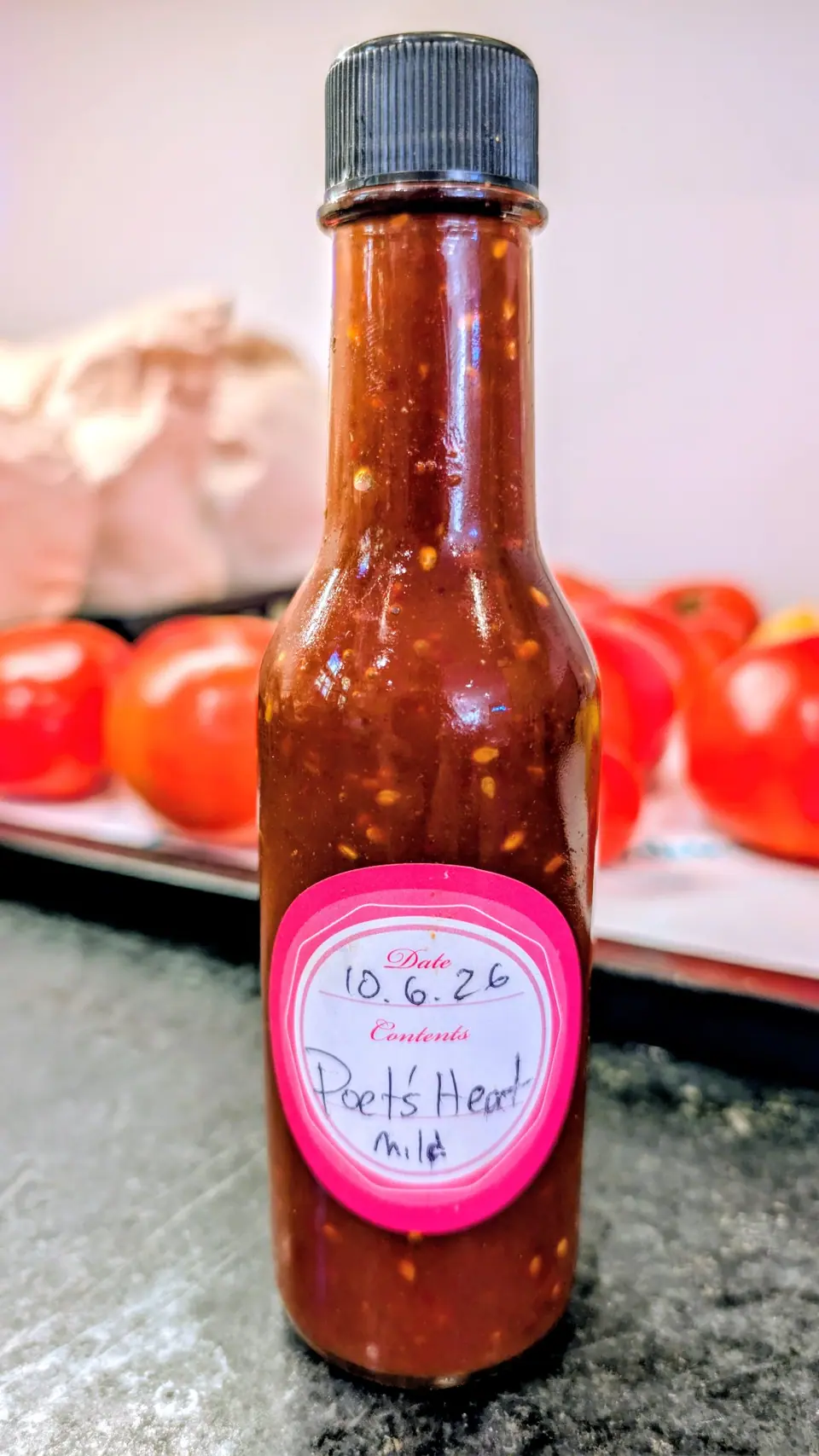 A narrow woozy bottle of thick brick-red sauce stands on dark speckled stone on 8 October, opaque and shot through with pale seeds, a thin line of oil separated on its right shoulder; behind it, blurred round red fruits on a white tray in soft indoor light.