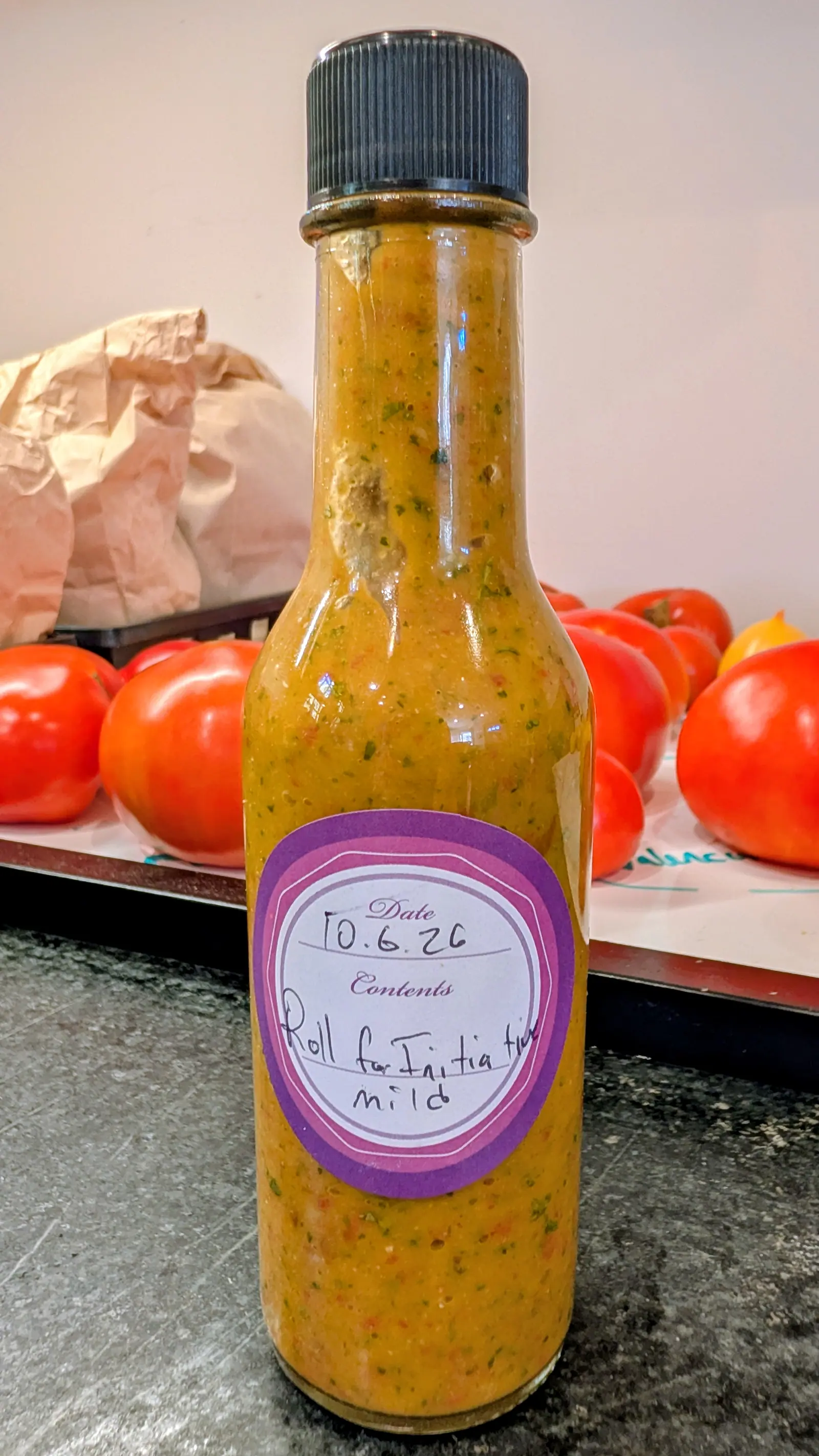 A tall narrow bottle of homemade sauce on 8 October, filled to the shoulder with opaque golden-orange puree flecked with green and red, a single clump hanging near the neck. Flat indoor light, glossy red fruit on a paper-lined tray behind.