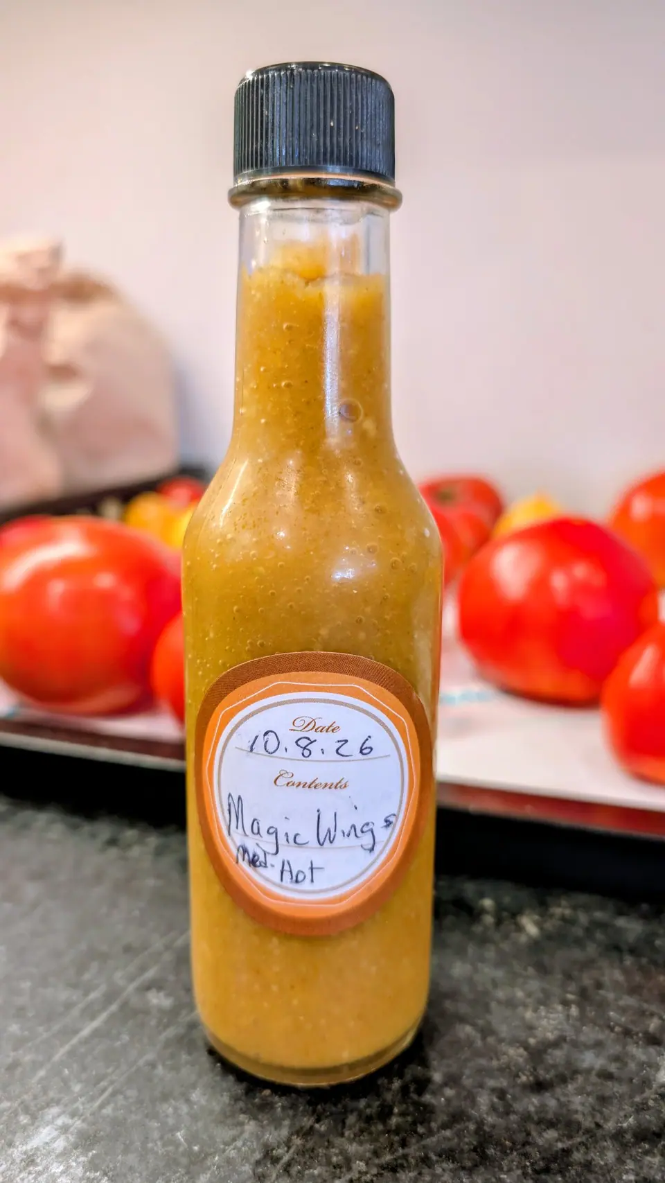 A slim woozy bottle of thick golden-orange puree stands on a dark speckled counter on 8 October, filled to the shoulder with pale flecks and small seeds suspended throughout and fine bubbles against the glass. Soft indoor light, no shadows.