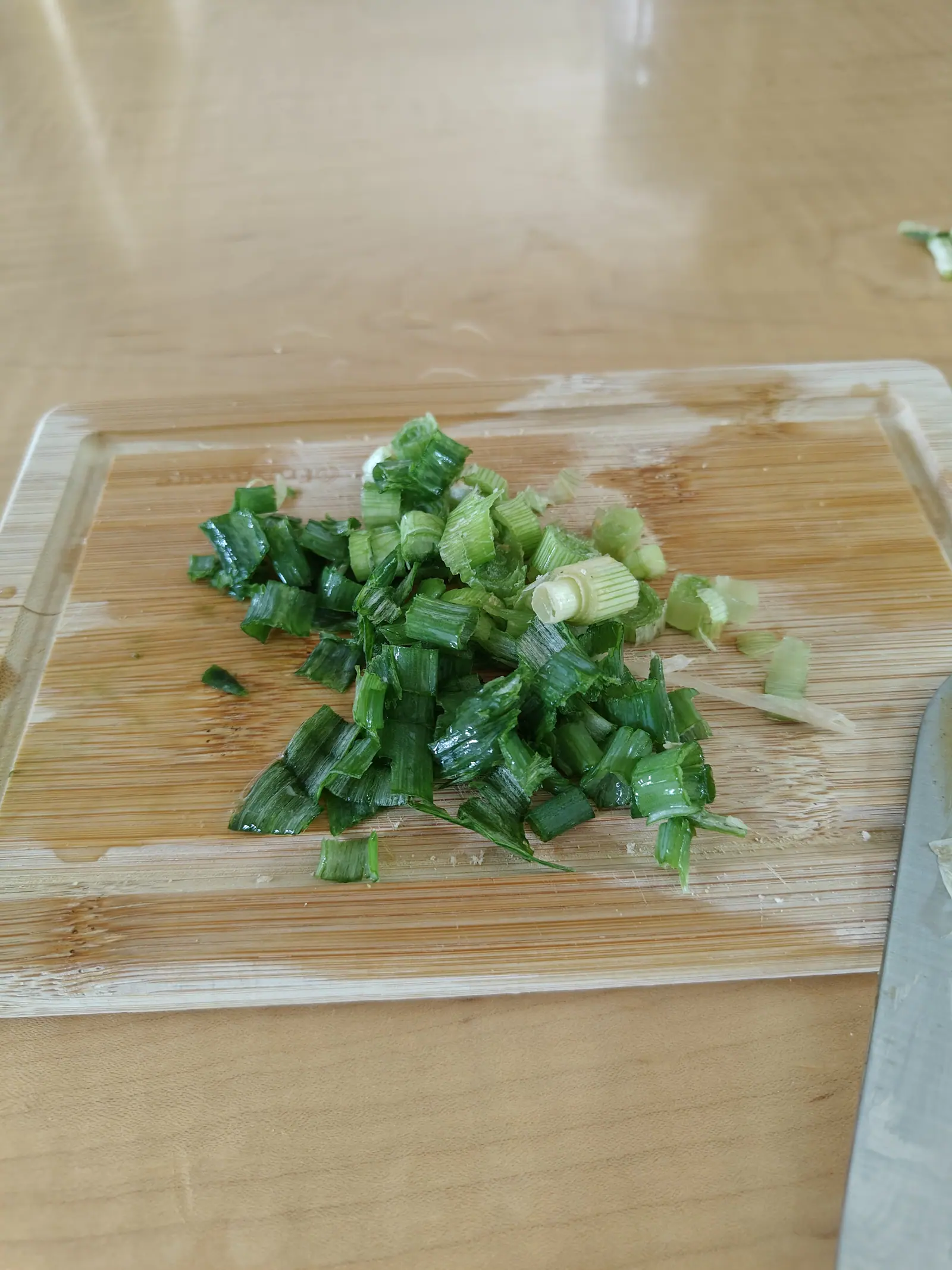 Chopped green stalk heaped on a bamboo board on 12 June, every piece cut across to show it hollow, and one pale cream segment near the middle opened to two rings nested inside each other, wet and glossy in soft daylight from the left.
