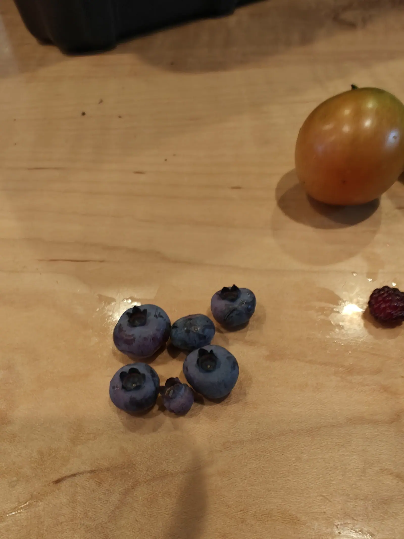 High Bush blueberries on 29 June, six of them loose on bare wood with a dark crown pricked into every upper face, five keeping their waxy bloom and one rubbed clean of it so that it reads darker and glossier than the rest in warm light.