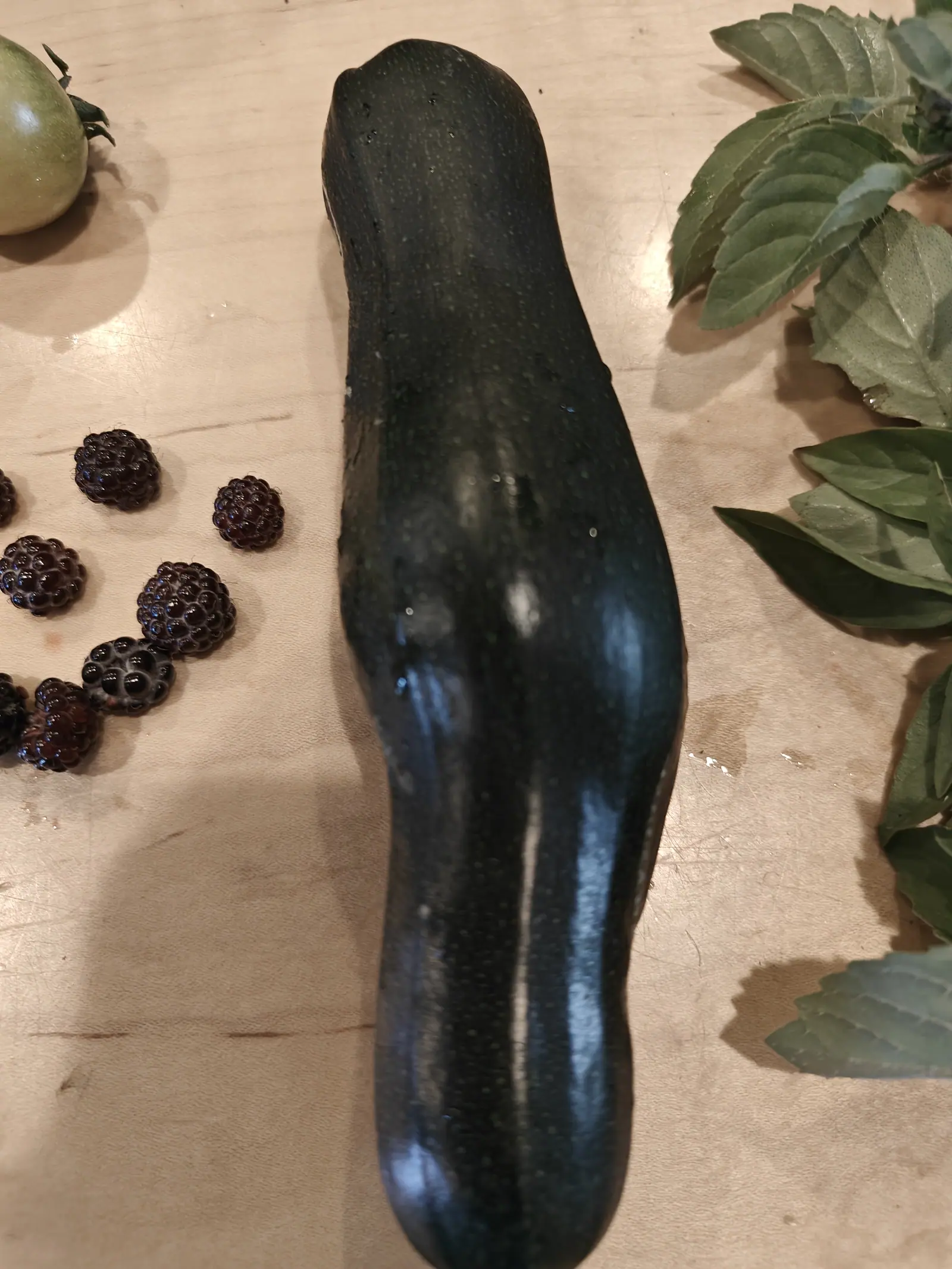 A zucchini washed and laid on the cutting board on 29 June, its near-black skin flecked all over with tiny pale green dots and still holding beads of water down one flank, a scatter of dark berries and one green tomato alongside.
