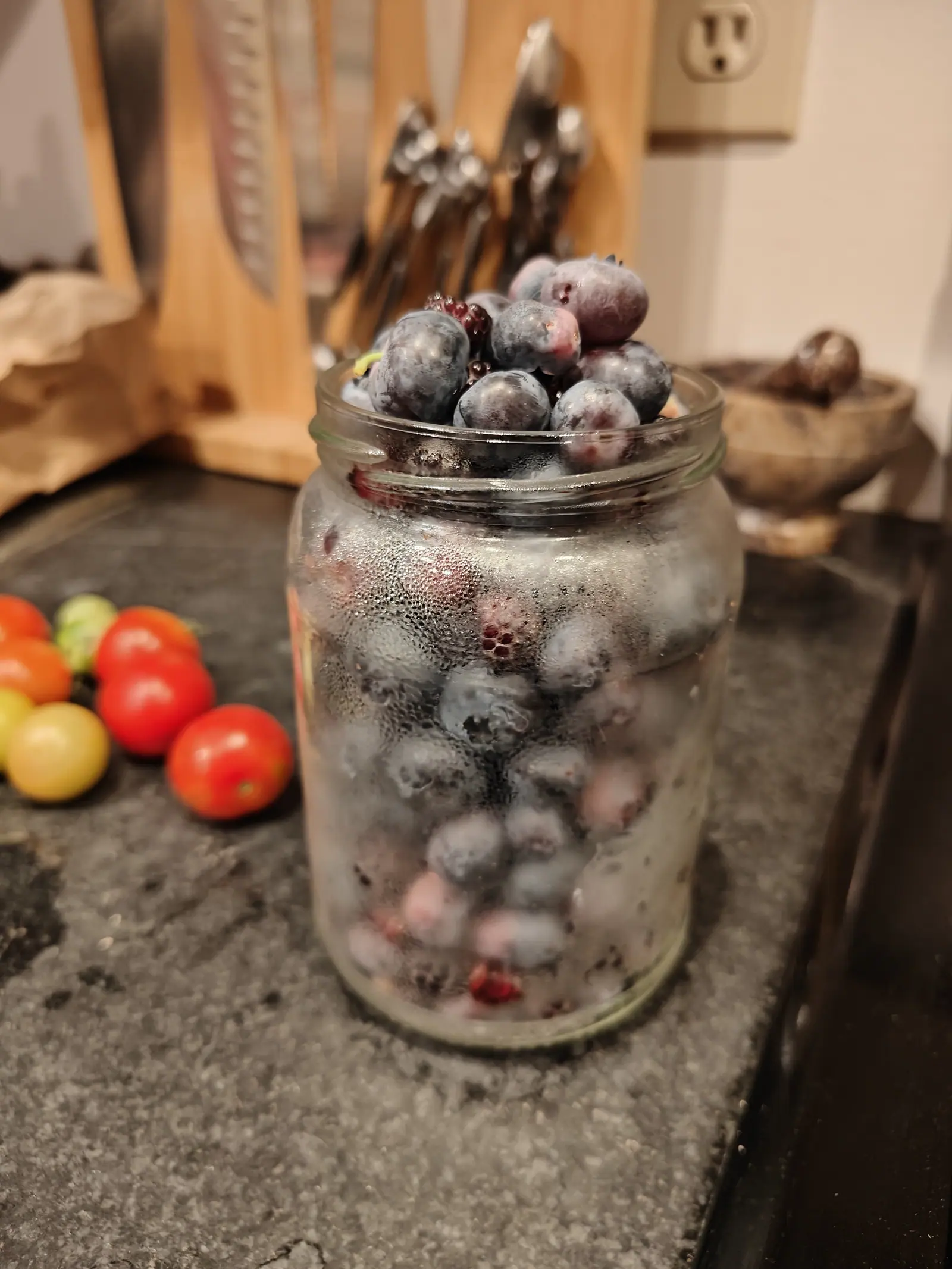 High Bush blueberries on 2 July heaped above the rim of a jar just out of the cold, the glass fogged right over so everything below the neck reads as blurred purple behind beaded water, their bloom smeared gray where hands went through them.