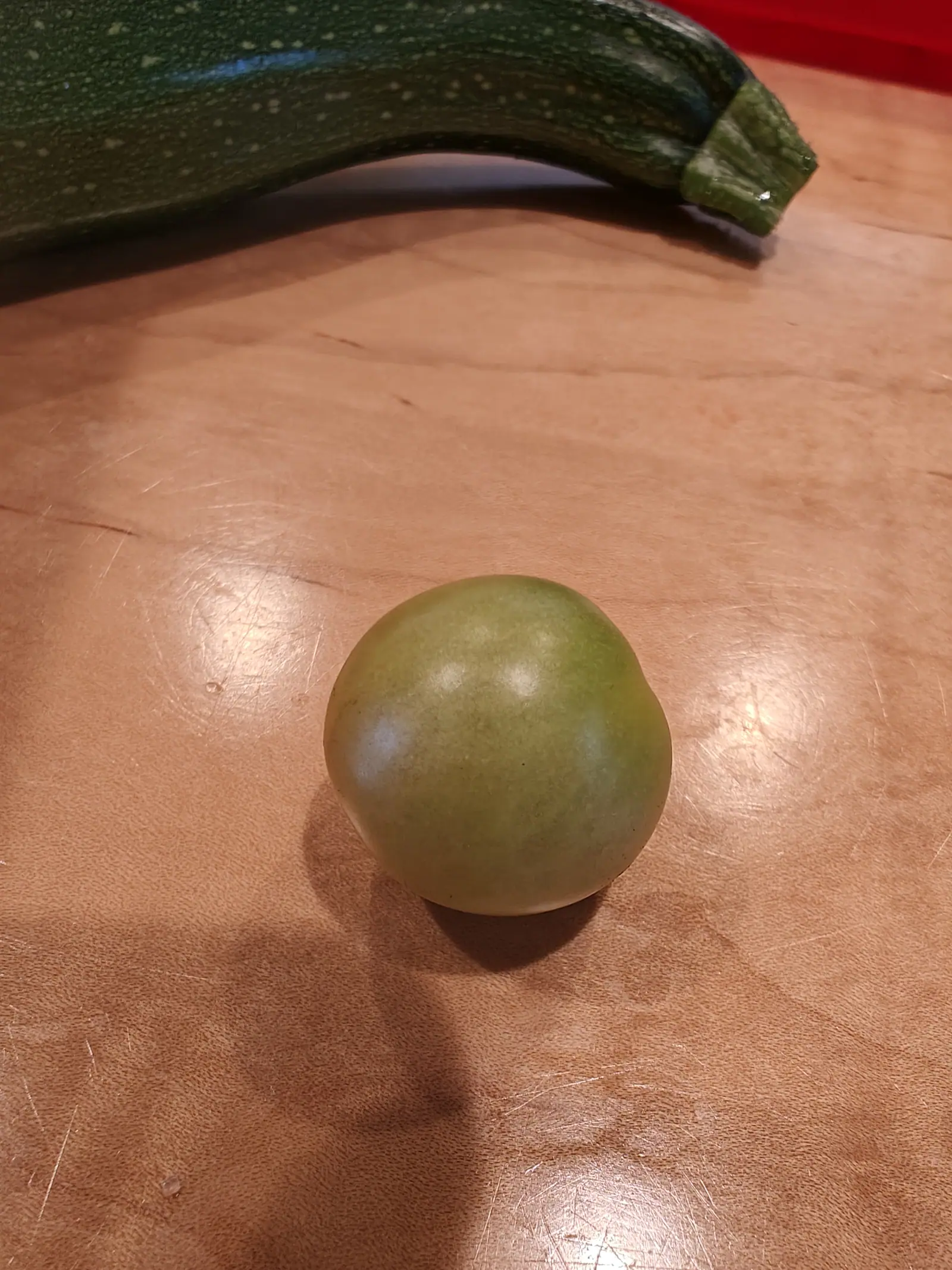 One round fruit on 12 July, picked and set on a scratched wooden board indoors — matt gray-green shading to creamy yellow across its lower half, no stalk or calyx left on it — with a long curved summer squash behind, flecked cream and faintly striped.