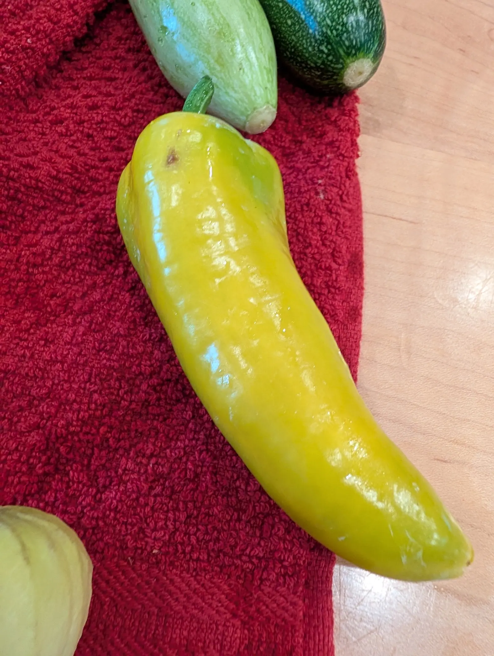 A Pick-N-Pop Yellow pepper picked on 16 July, clear yellow through the body but still washed pale green on one shoulder and again at the very tip, three shallow creases down its near side, laid across a deep red towel under flat kitchen light.